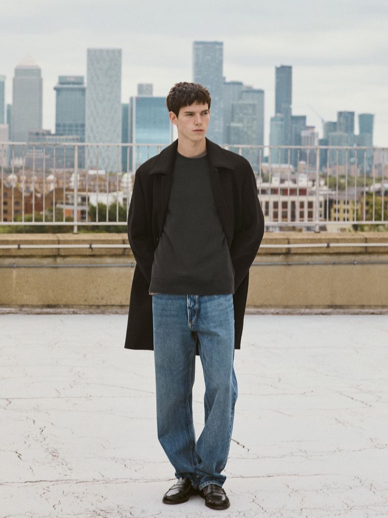 Young man wearing a black coat, grey t-shirt, and blue jeans standing on a rooftop with a city skyline in the background.
