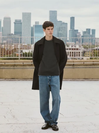 Young man wearing a black coat, grey t-shirt, and blue jeans standing on a rooftop with a city skyline in the background.