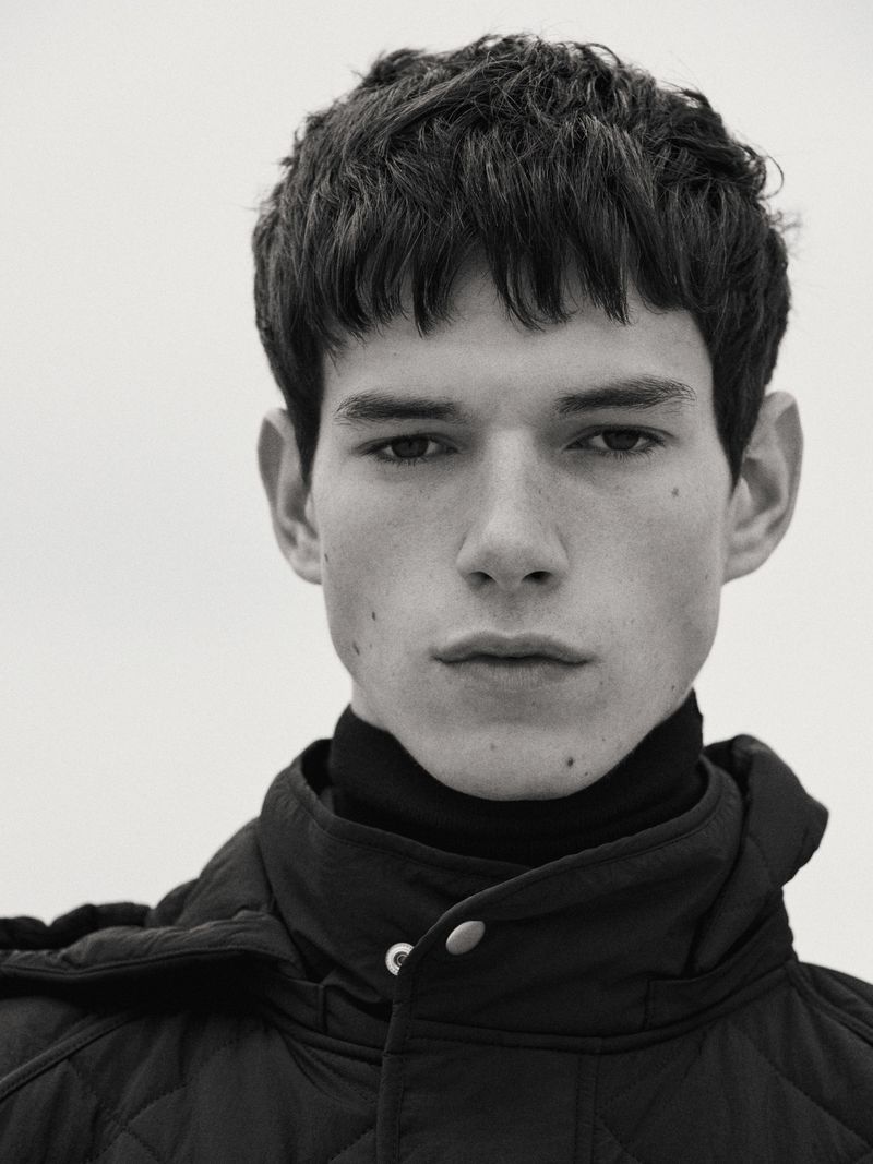 Monochrome portrait of a young adult male with short brown hair wearing a black quilted jacket and turtleneck against a plain background.