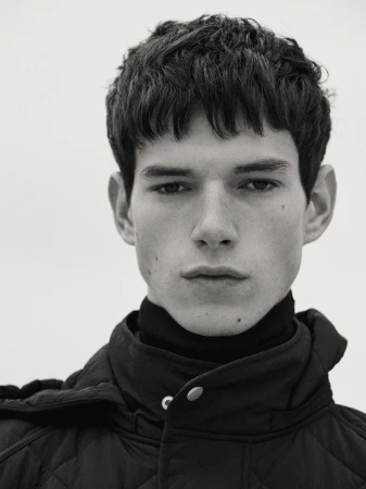 Monochrome portrait of a young adult male with short brown hair wearing a black quilted jacket and turtleneck against a plain background.