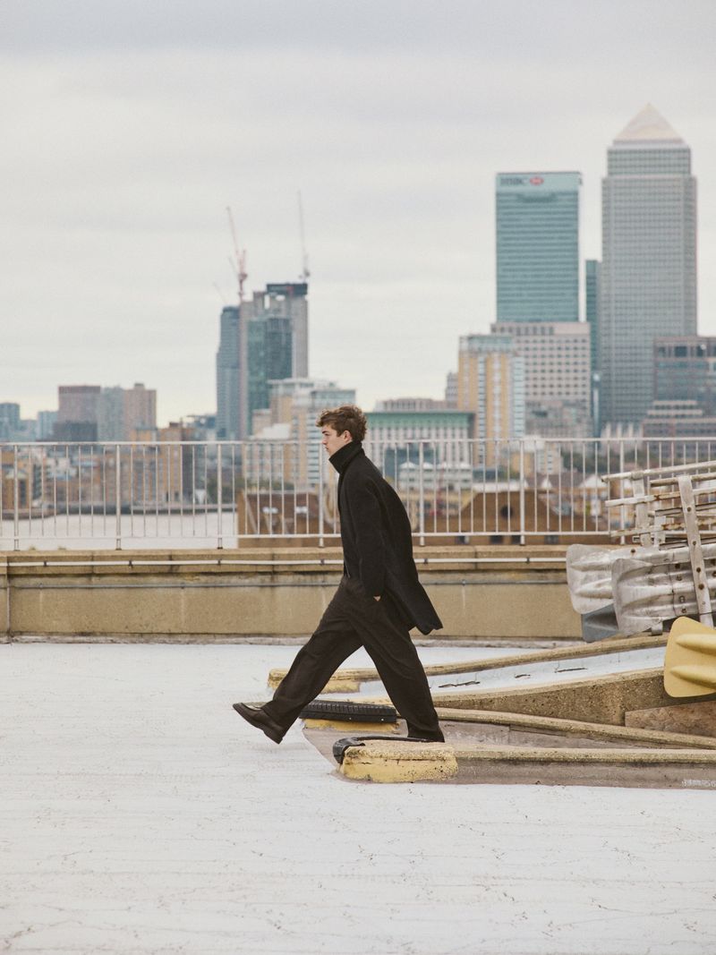 A young adult man in a black overcoat walks on a rooftop in front of a city skyline with modern buildings on an overcast day.