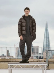 A young male model standing on a rooftop wearing a dark brown puffer jacket, grey shirt, and trousers against a city skyline.
