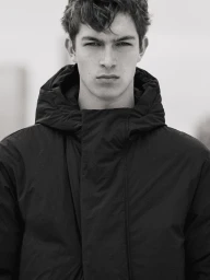Close-up portrait of a male model wearing a dark hooded puffer jacket against a neutral backdrop.