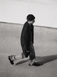 A young male model walking outdoors wearing a black overcoat, denim jeans, and dark shoes against a textured urban background.