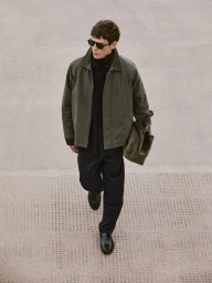 Model in olive green jacket and dark trousers walking outdoors, carrying a matching leather bag.