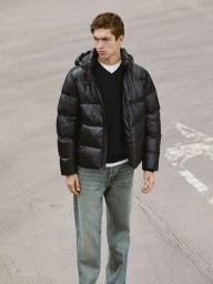 Model wearing a black puffer jacket and blue jeans standing outdoors against a grey asphalt pavement background.