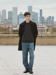 Young man wearing a black coat, grey t-shirt, and blue jeans standing on a rooftop with a city skyline in the background.