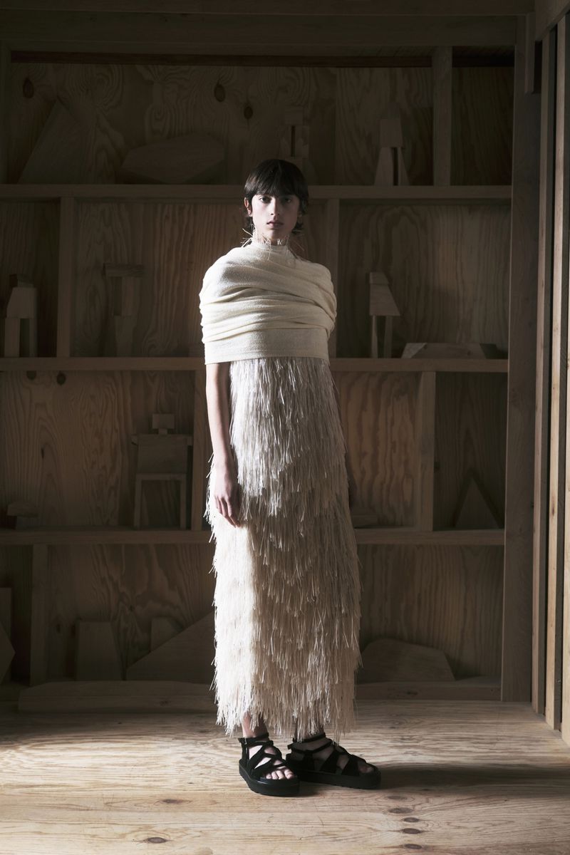 A woman in a long fringe dress stands in an art gallery-like space with light wood walls and shelves holding sculptures.