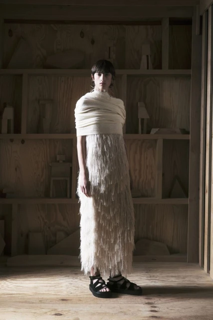 A woman in a long fringe dress stands in an art gallery-like space with light wood walls and shelves holding sculptures.
