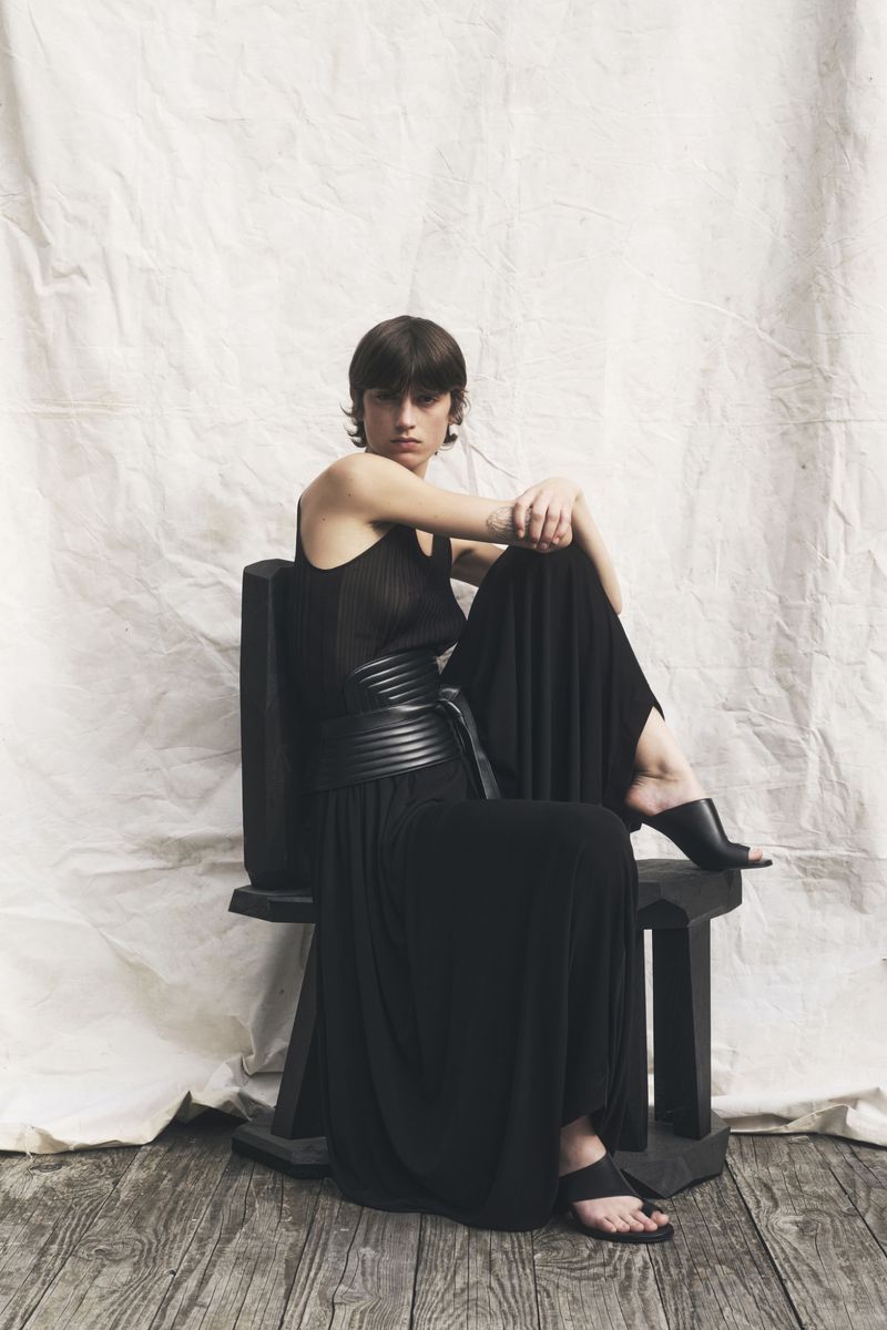 A woman in a black dress sits on a wooden chair in a studio, exuding relaxed confidence in a fashion-forward editorial setting.