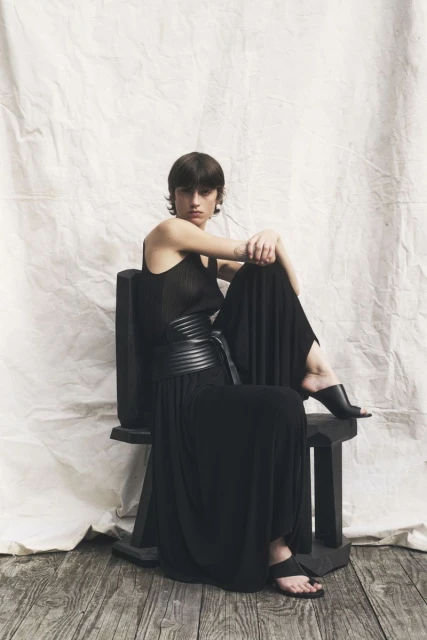 A woman in a black dress sits on a wooden chair in a studio, exuding relaxed confidence in a fashion-forward editorial setting.