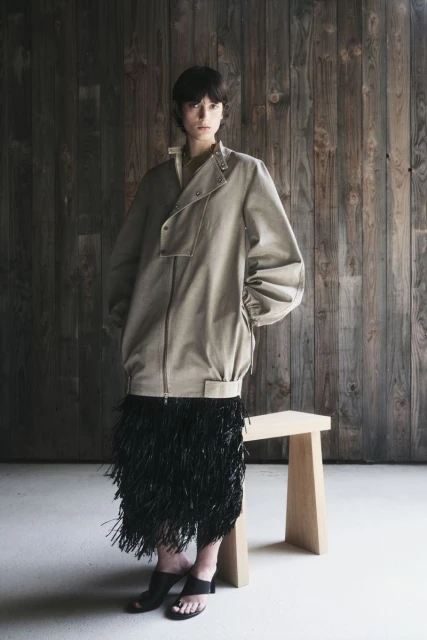 A woman poses in a beige zipper jacket and black fringe skirt in a studio with a wooden backdrop and a modern, light-wood stool.