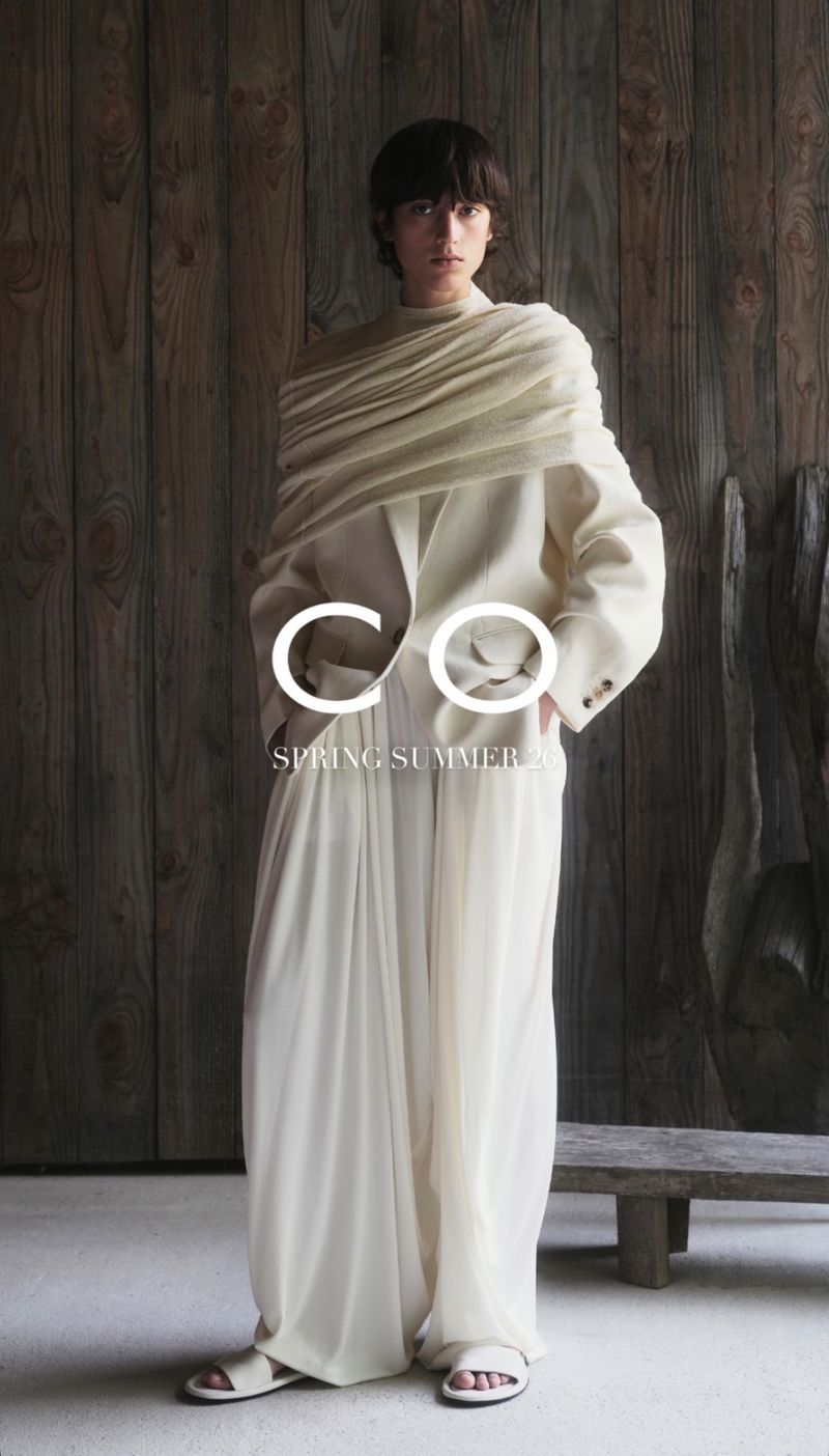 An adult female fashion model poses in a full-length studio shot wearing an off-white suit with a draped shawl and strappy sandals.