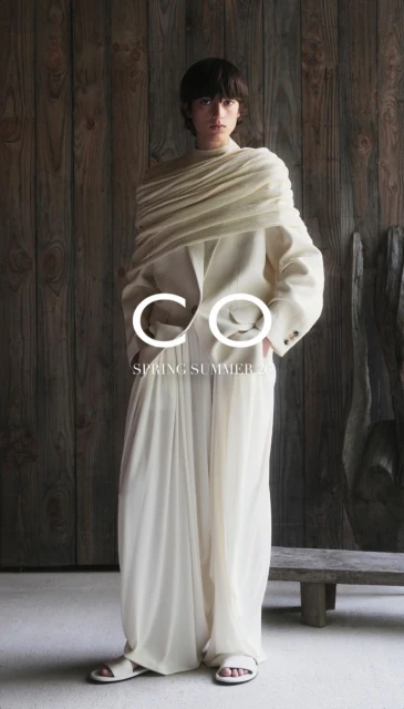 An adult female fashion model poses in a full-length studio shot wearing an off-white suit with a draped shawl and strappy sandals.