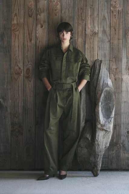 A woman in an olive jumpsuit stands in front of a weathered wooden wall with a driftwood sculpture, showcasing contemporary fashion.