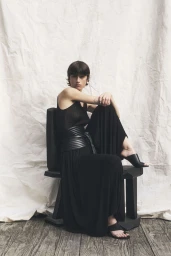 Model in a black outfit sitting on a dark chair against a textured backdrop for the CO lookbook SS26.