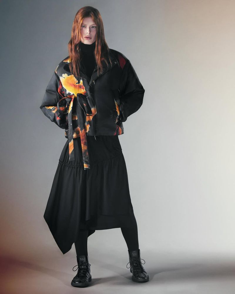 Fashionable redhead woman in a black turtleneck, printed jacket, and asymmetrical skirt poses in a studio with a neutral background.