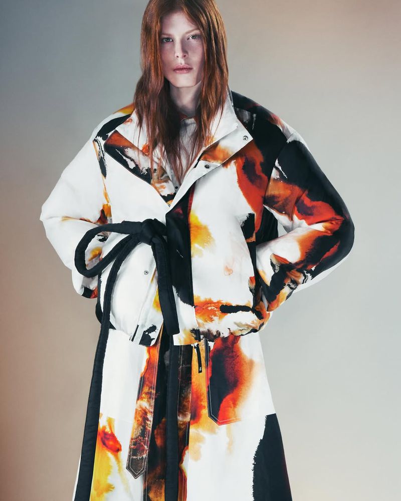 A person poses in a studio wearing a jacket and skirt with abstract orange and black designs, accented by a tied black rope belt.