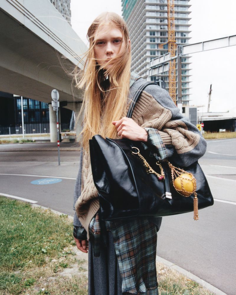Fashion shot of a young adult woman with long blonde hair, wearing a sweater and skirt and carrying a large black leather bag outdoors.