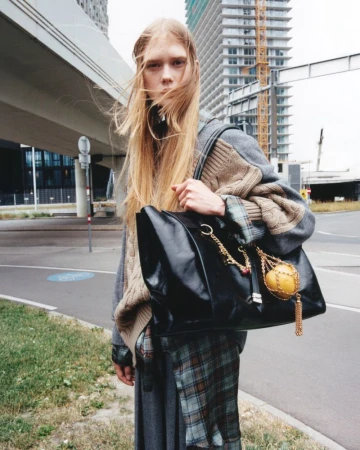 Fashion shot of a young adult woman with long blonde hair, wearing a sweater and skirt and carrying a large black leather bag outdoors.