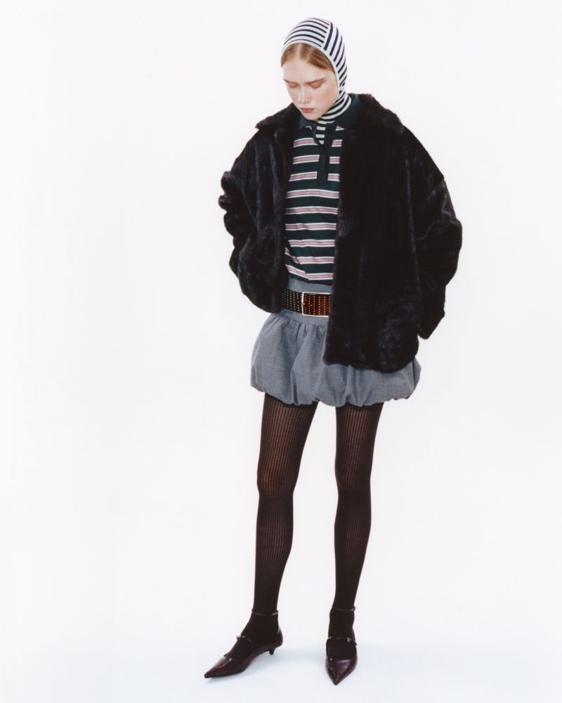 Fashionable woman in faux fur coat, striped knit shirt and beanie, puffy skirt, tights, and buckle shoes poses in studio.