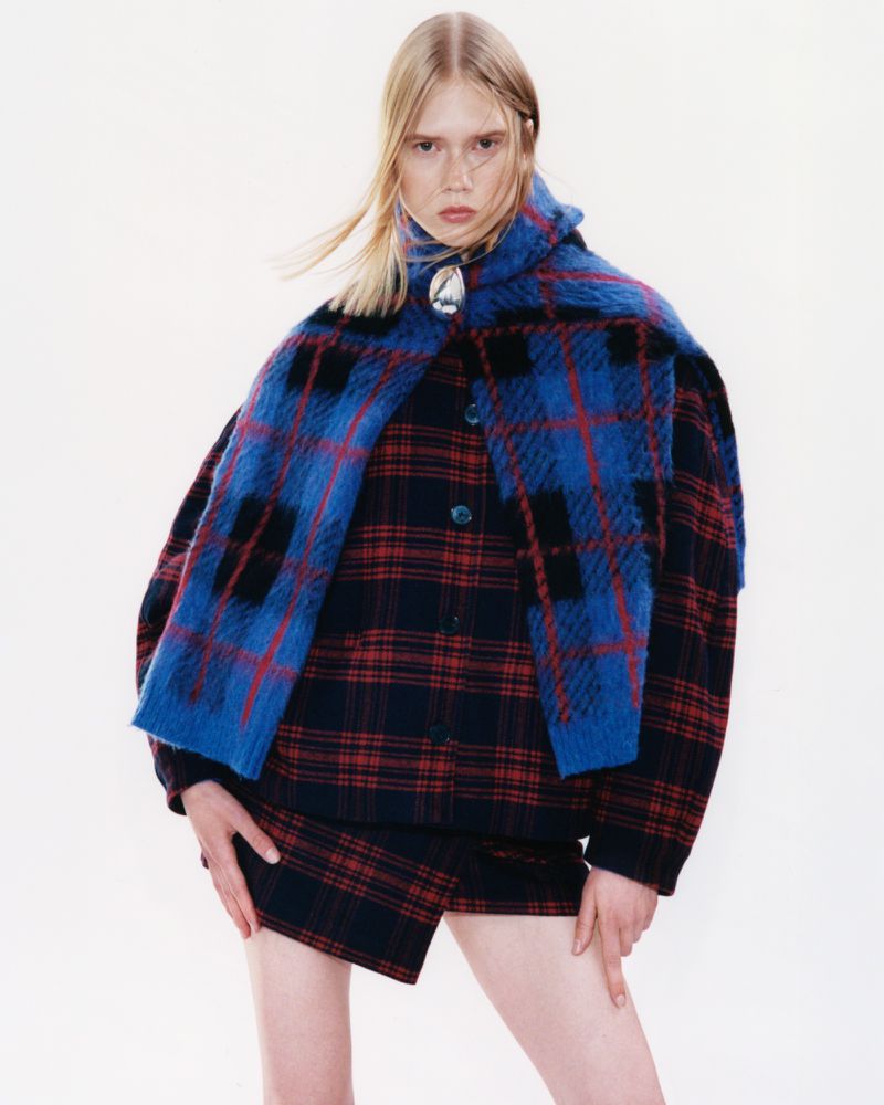 Fashion shot of a blond model wearing a blue and red plaid outfit and cape against a bright white background in a studio setting.