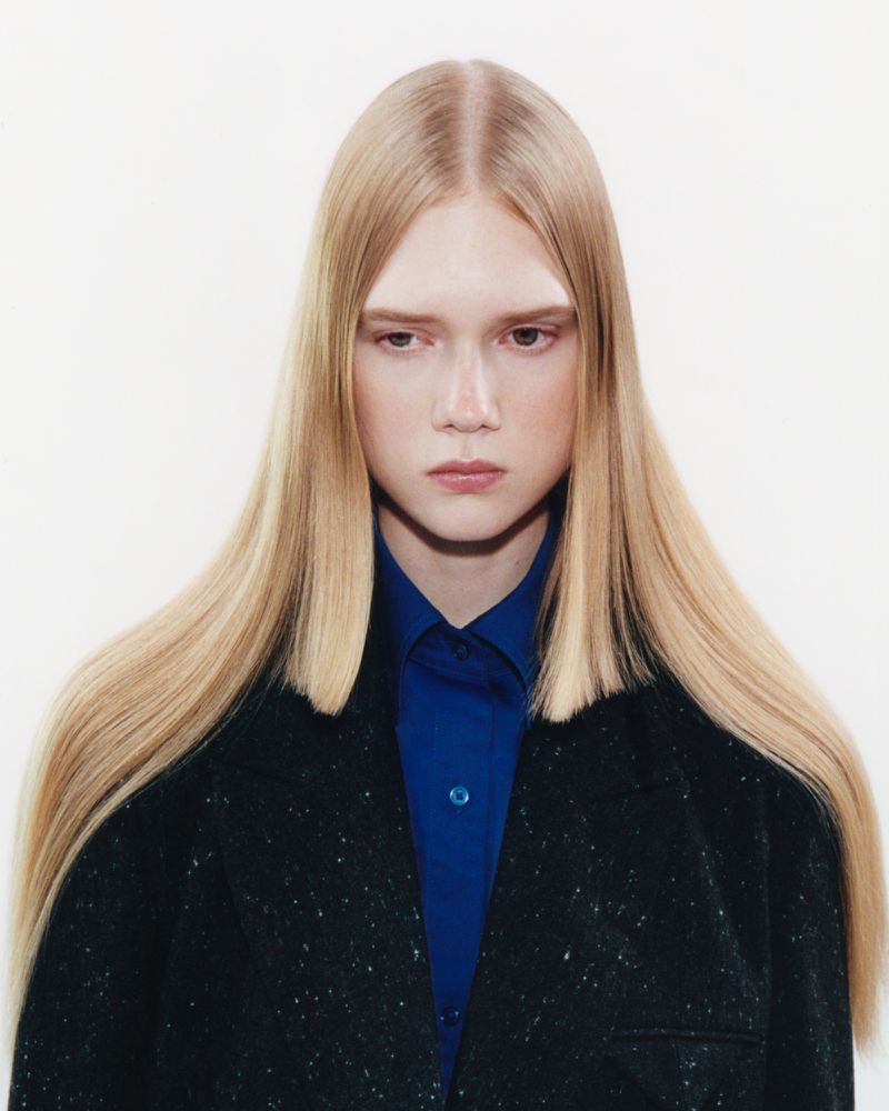 Model with straight blonde hair wearing a blue button-down shirt and black jacket against a plain white background
