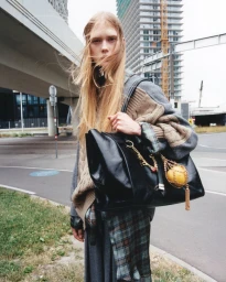Model standing outdoors near a roadway holding a large black leather handbag with gold chain charms for EDITED.de.
