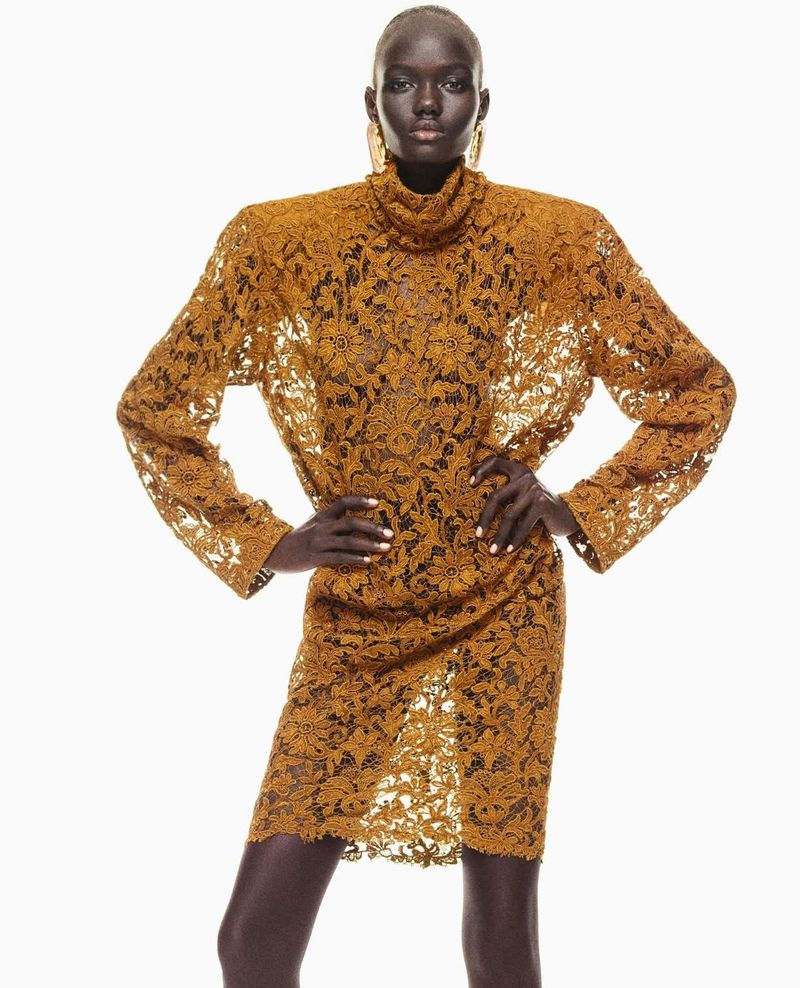 Portrait of a Black female model wearing a gold floral-lace turtleneck dress, posing against a white backdrop in a fashion editorial shot.