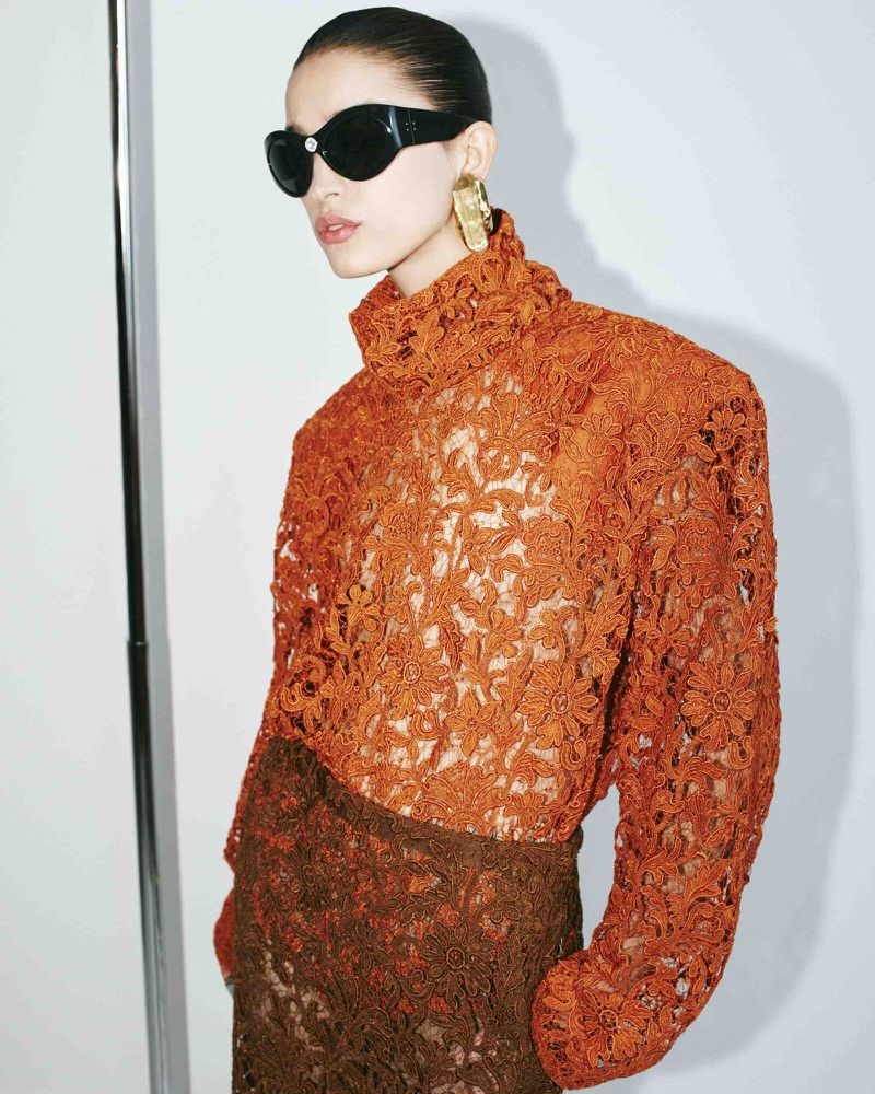 A woman in an orange lace top and sunglasses poses in front of a white wall, showcasing fashion and style with a modern, artistic flair.