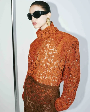 A woman in an orange lace top and sunglasses poses in front of a white wall, showcasing fashion and style with a modern, artistic flair.