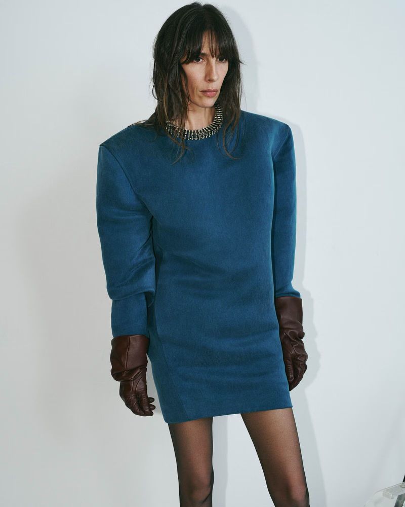 Studio shot of a brunette woman in a blue mini dress and brown gloves, posing against a white backdrop with neutral expression.