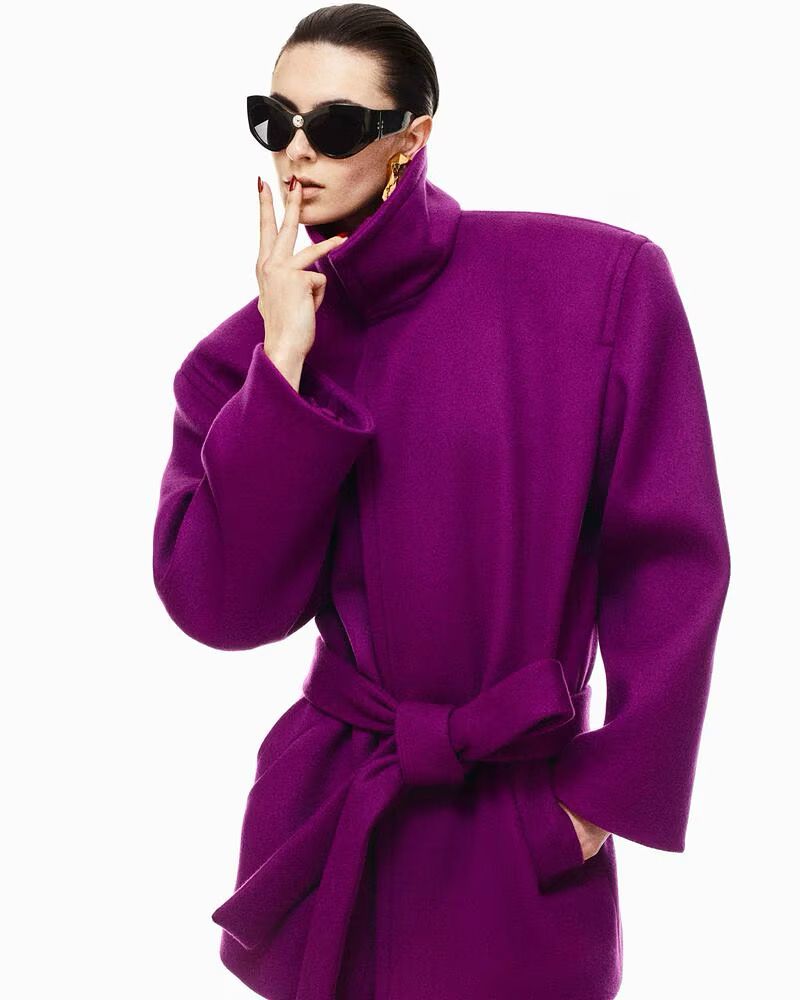 Woman in a purple coat and black sunglasses poses against a white backdrop with hands near her face and gold earrings.