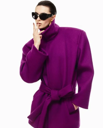 Woman in a purple coat and black sunglasses poses against a white backdrop with hands near her face and gold earrings.