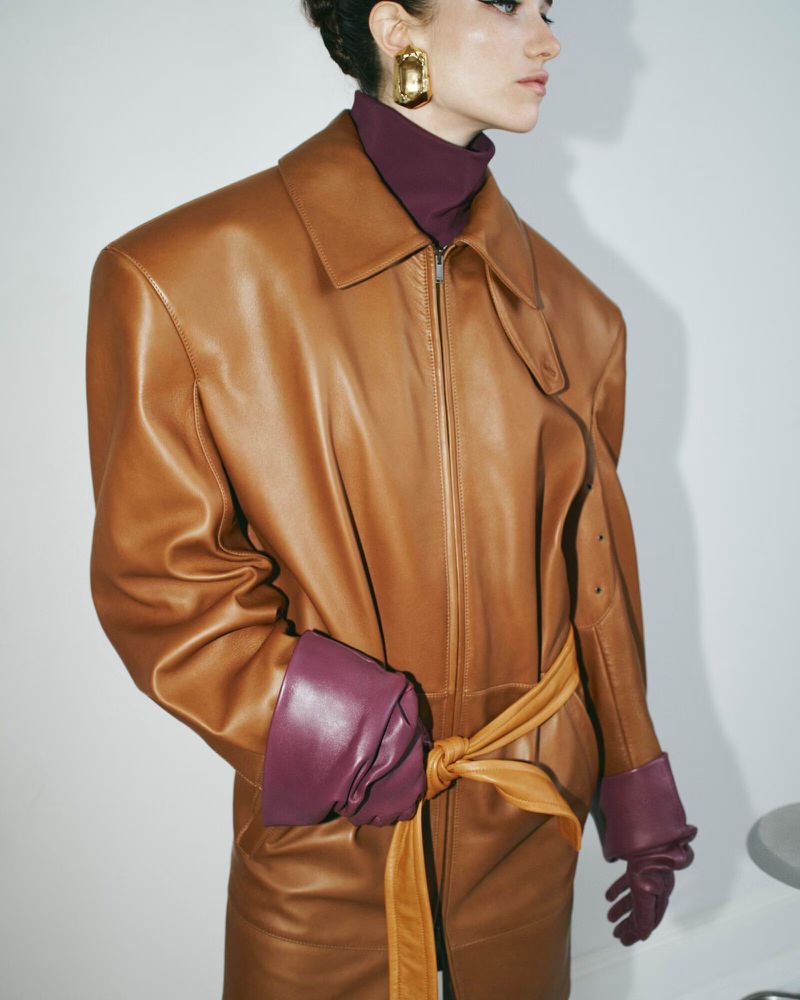 Woman poses in tan leather coat with maroon gloves and turtleneck against a white backdrop, showcasing fashion and style.