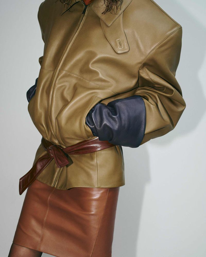A woman showcases a stylish ensemble featuring a tan leather bomber jacket cinched with a brown belt, paired with a matching leather skirt.