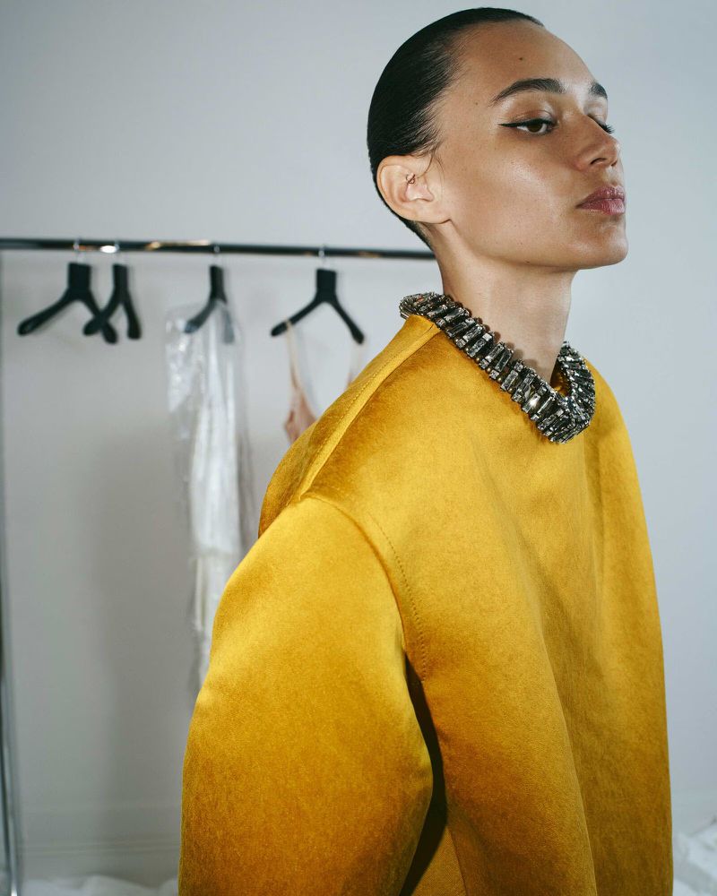 Elegant adult female poses in mustard yellow satin top and jeweled collar. Hair slicked back, clean background. Fashion, beauty, and portrait.