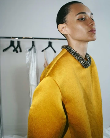 Elegant adult female poses in mustard yellow satin top and jeweled collar. Hair slicked back, clean background. Fashion, beauty, and portrait.