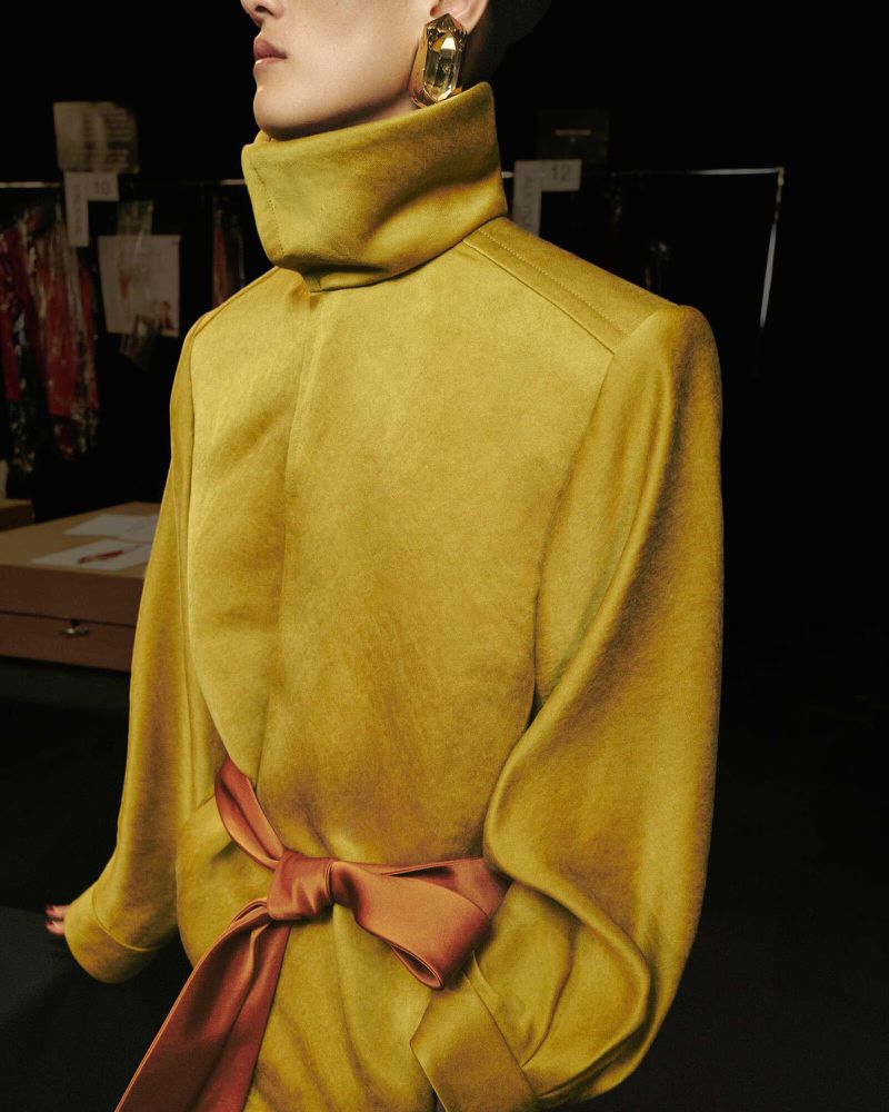 Fashion shot of a person wearing a yellow high-neck top with a satin bow, gold earrings, against a black background. Studio lighting.