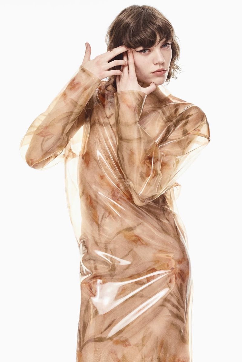 Woman in sheer floral-patterned raincoat poses in front of white background. Fashion-forward pose with hands near face.