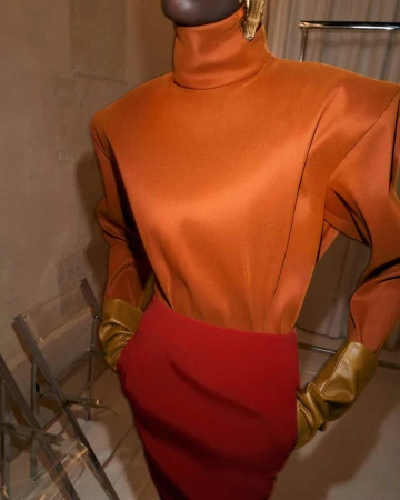 A dark-skinned woman wearing an orange high-necked blouse with shoulder pads and long brown gloves poses with a red pencil skirt.