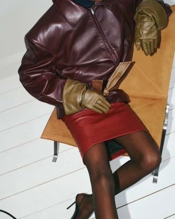 Fashion shot: Adult female poses seated wearing a leather jacket, red skirt, tights, and high heels in an indoor setting.