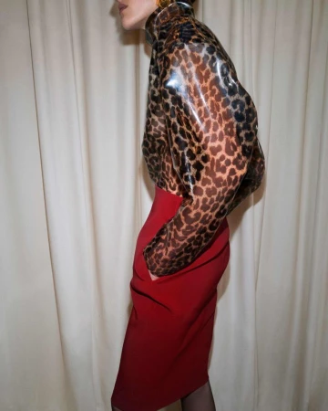 Adult female fashion model poses in a leopard print jacket and red skirt against a tan fabric backdrop in a medium studio shot.