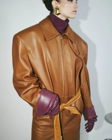 Model wearing a tan leather coat with a matching tie belt, purple gloves, and a purple turtleneck against a plain white background
