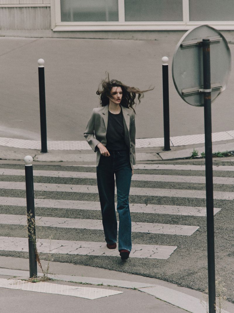 A woman in casual fashion strides across a zebra-striped crosswalk, her hair blowing, in a neutral-toned urban setting with geometric architecture.