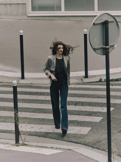 A woman in casual fashion strides across a zebra-striped crosswalk, her hair blowing, in a neutral-toned urban setting with geometric architecture.