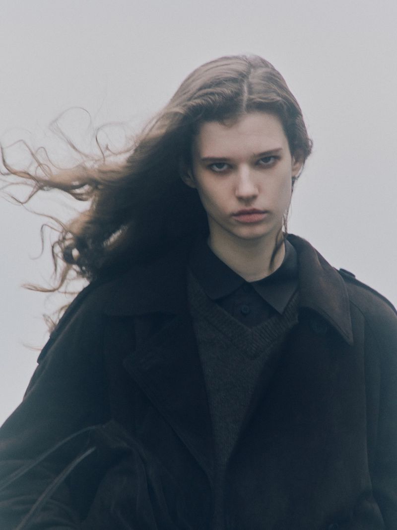 Portrait of a young adult woman with long brown hair and a dark coat, standing outside against a light grey sky in overcast weather.
