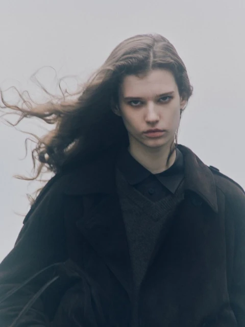 Portrait of a young adult woman with long brown hair and a dark coat, standing outside against a light grey sky in overcast weather.