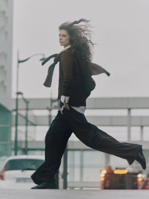 A stylish woman with long hair strides confidently across an urban street in a fashion editorial shot, wearing a brown sweater and loose pants.
