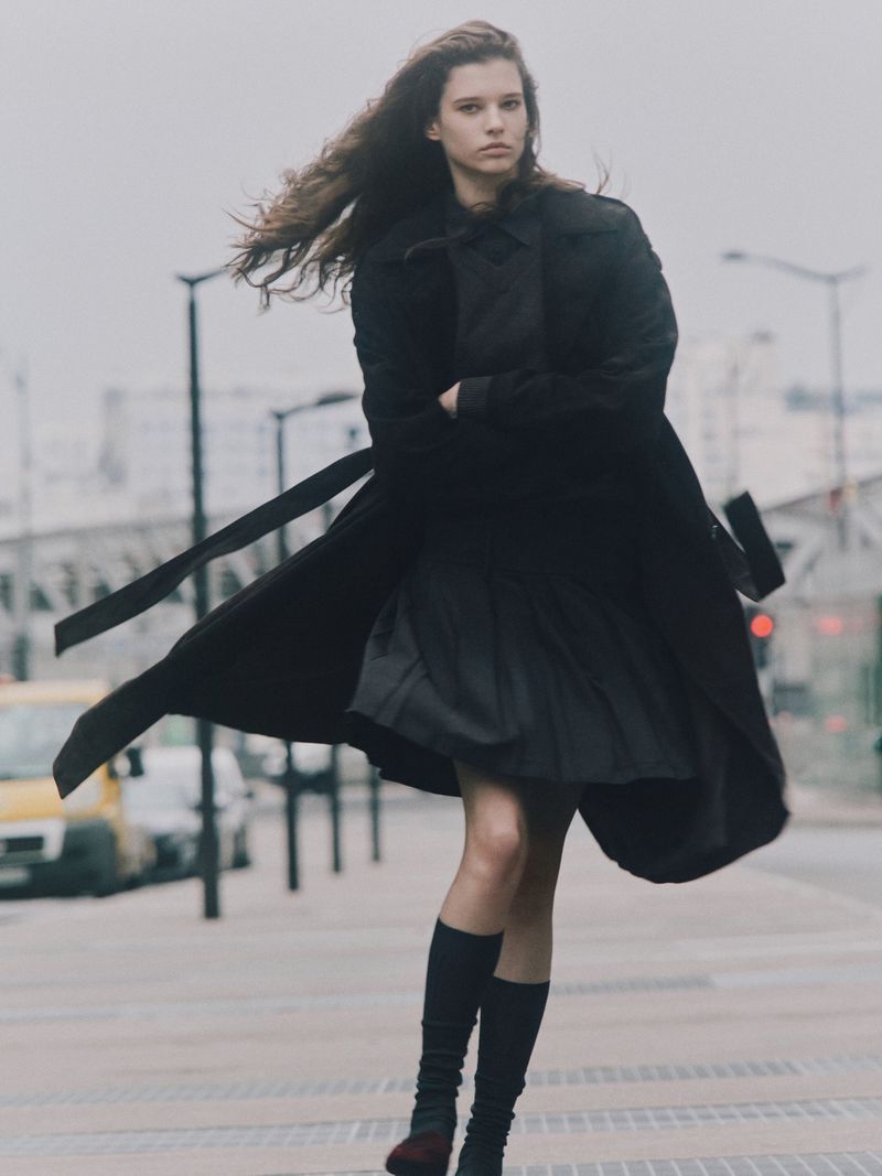 A young woman in a dark coat and knee-high socks poses on a city street in overcast weather, with a blurry vehicle in the background.